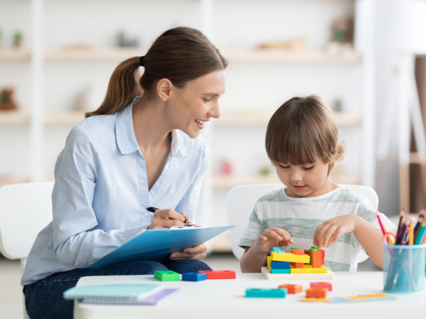 Early kids development. Cute little boy playing logical geometry game, positive woman specialist with notepad watching him at office
