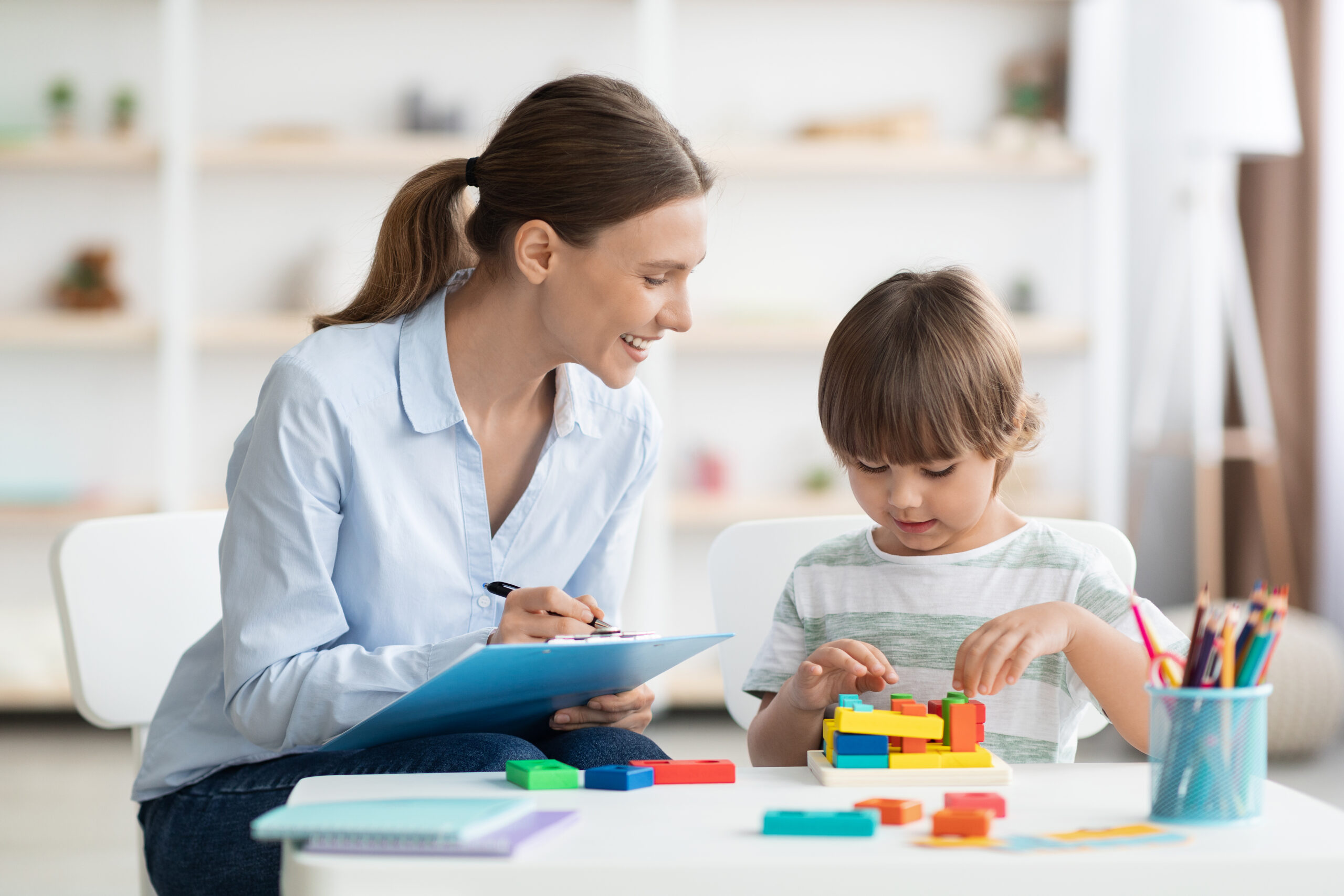 Early kids development. Cute little boy playing logical geometry game, positive woman specialist with notepad watching him at office